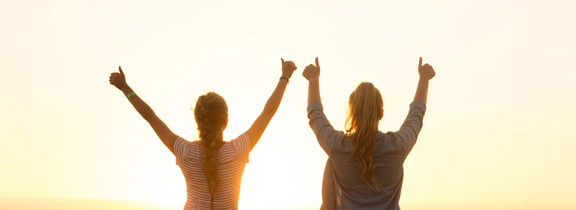 Zwei Mädchen stehen mit erhobenen Händen vorm Sonnenuntergang