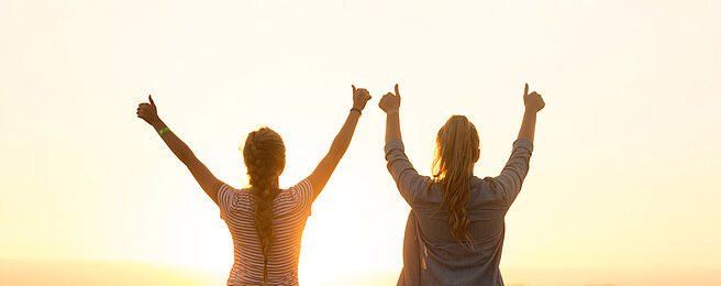 Zwei Mädchen stehen mit erhobenen Händen vorm Sonnenuntergang