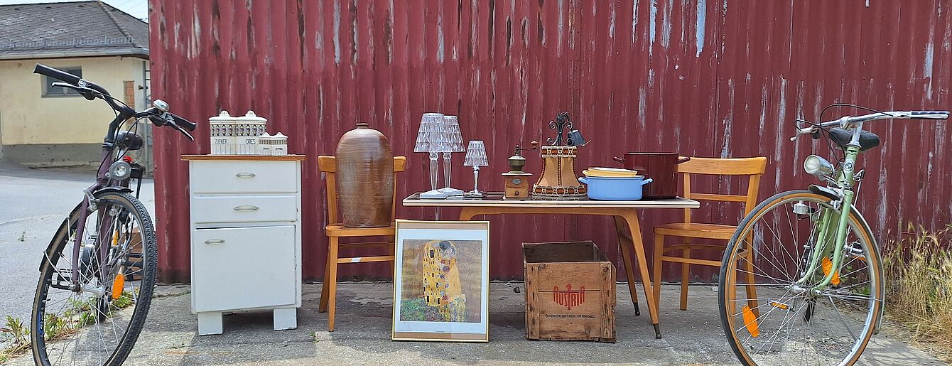 Foto: Lilly Kolb Zwei Fahrräder, ein weißer Nachttisch, ein Tisch mit zwei Stühlen, Lampen, ein Bild und eine Holzkiste vor einer roten Holzwand