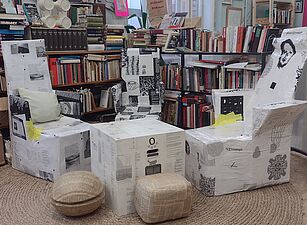 Sessel und Hocker aus Karton mit schwarz-weißen Aufdrucken in einem Raum mit Bücherregalen und einer Person im Hintergrund.