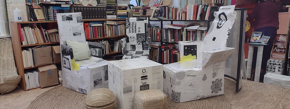 Sessel und Hocker aus Karton mit schwarz-weißen Aufdrucken in einem Raum mit Bücherregalen und einer Person im Hintergrund.