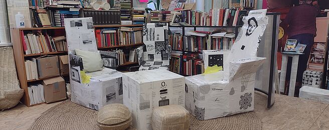 Sessel und Hocker aus Karton mit schwarz-weißen Aufdrucken in einem Raum mit Bücherregalen und einer Person im Hintergrund.