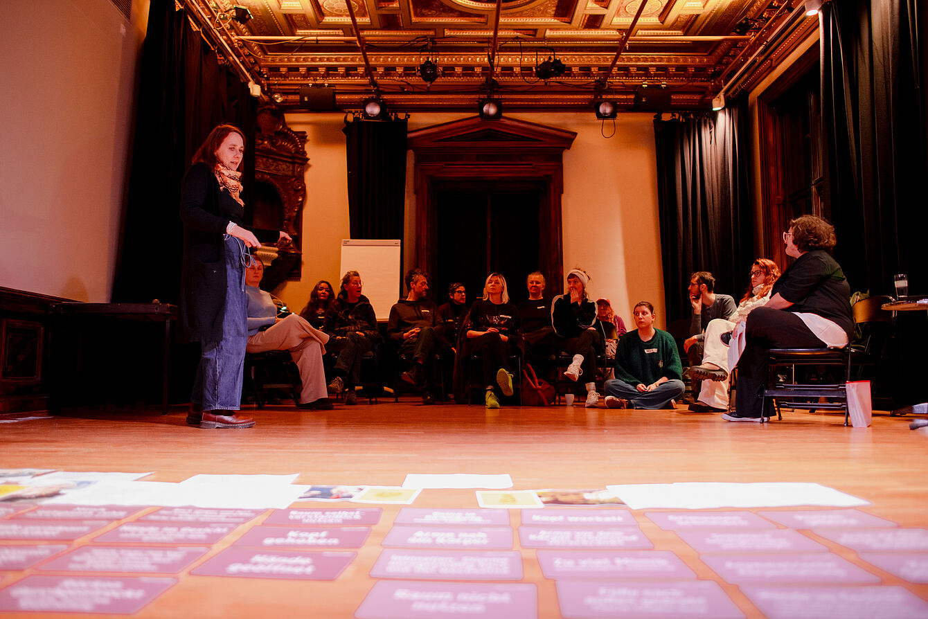 Gruppe von Menschen sitzt und steht in einem großen Raum mit Holzdecke, einige sitzen auf Stühlen, andere auf dem Boden, im Vordergrund liegen Karten und Zettel auf dem Boden.