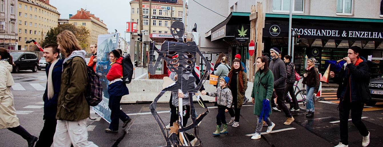 Pedestrian crossing with people walking past a large skeleton figure holding a sign on a city street corner.