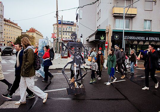 Große Skelettfigur aus Pappe auf einem Rollbrett, umgeben von Fußgänger*innen auf einer Straßenkreuzung in der Stadt.