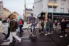 Große Skelettfigur aus Pappe auf einem Rollbrett, umgeben von Fußgänger*innen auf einer Straßenkreuzung in der Stadt.