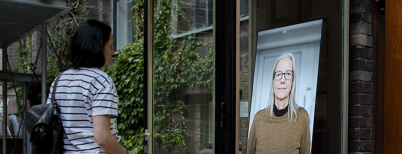 Person mit Rucksack und gestreiftem T-Shirt steht vor der Kunstzelle, in dem Screen zu sehen ist auf dem eine Person mit langem, hellem Haar und braunem Pullover zu sehen ist.