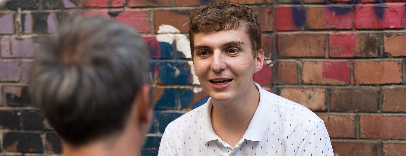 Jugendliche Person in weißem Poloshirt spricht mit älterer Person vor einer bemalten Backsteinwand