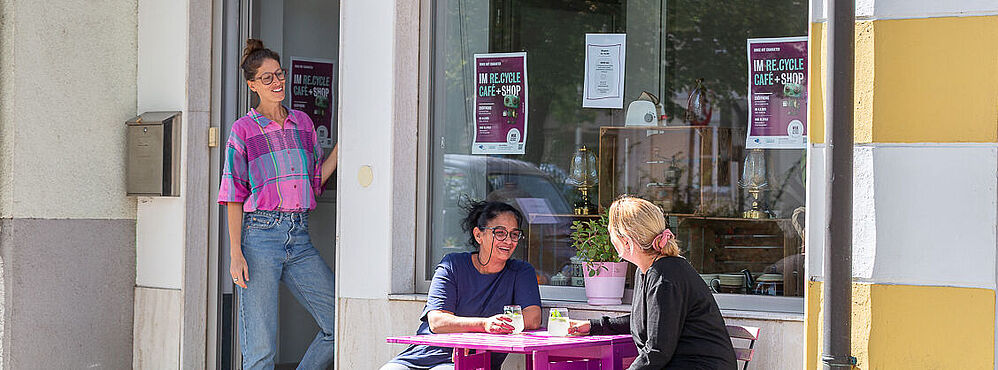 Foto: Susanne Senekowitsch wei Personen sitzen an einem kleinen lila Tisch vor einem Café, eine weitere Person steht in der Tür. Bunte Girlande hängt im Fenster