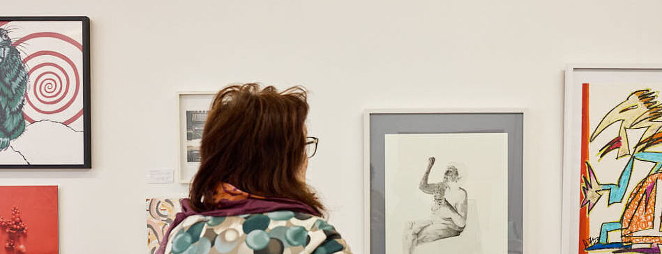 Eine Person mit Brille und gemustertem Pullover betrachtet gerahmte Kunstwerke an einer weißen Museumswand