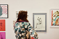 Eine Person mit Brille und gemustertem Pullover betrachtet gerahmte Kunstwerke an einer weißen Museumswand