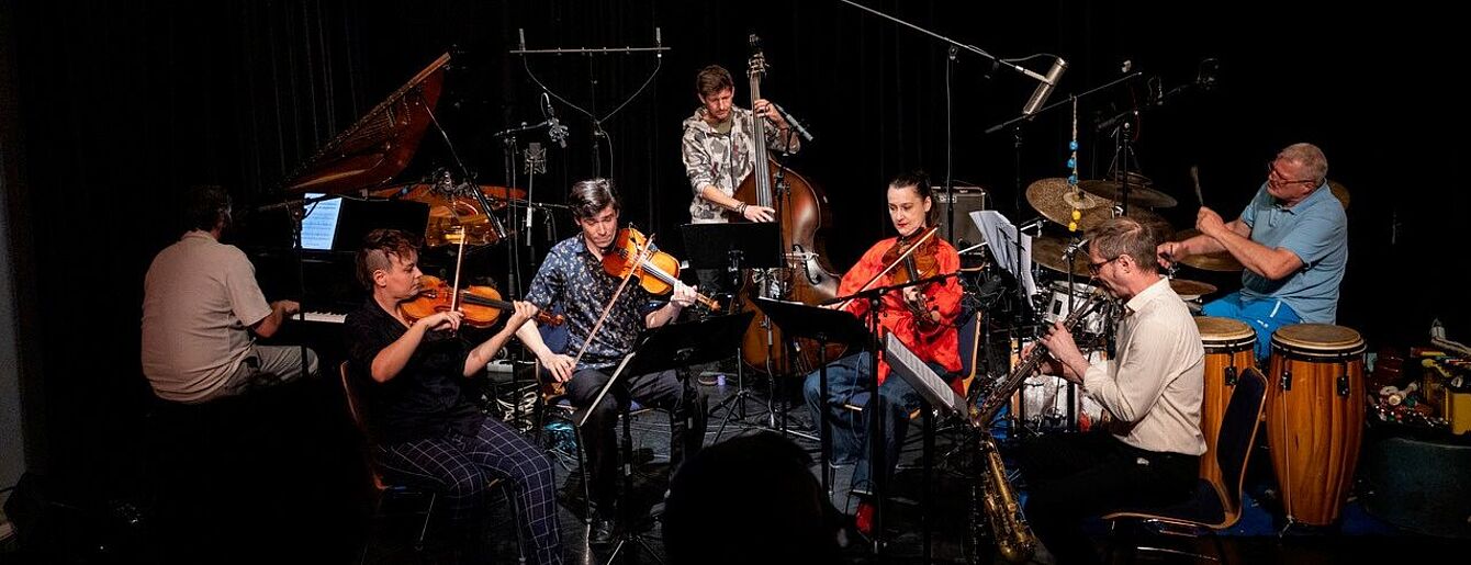 Sieben Musiker*innen spielen auf einer Bühne verschiedene Instrumente wie Klavier, Geige, Kontrabass, Saxophon und Schlagzeug in einem dunklen Raum