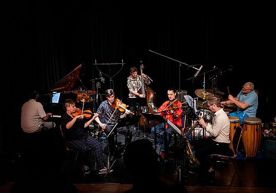 Sieben Musiker*innen spielen auf einer Bühne verschiedene Instrumente wie Klavier, Geige, Kontrabass, Saxophon und Schlagzeug in einem dunklen Raum