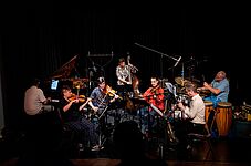 Sieben Musiker*innen spielen auf einer Bühne verschiedene Instrumente wie Klavier, Geige, Kontrabass, Saxophon und Schlagzeug in einem dunklen Raum