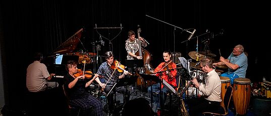 Sieben Musiker*innen spielen auf einer Bühne verschiedene Instrumente wie Klavier, Geige, Kontrabass, Saxophon und Schlagzeug in einem dunklen Raum