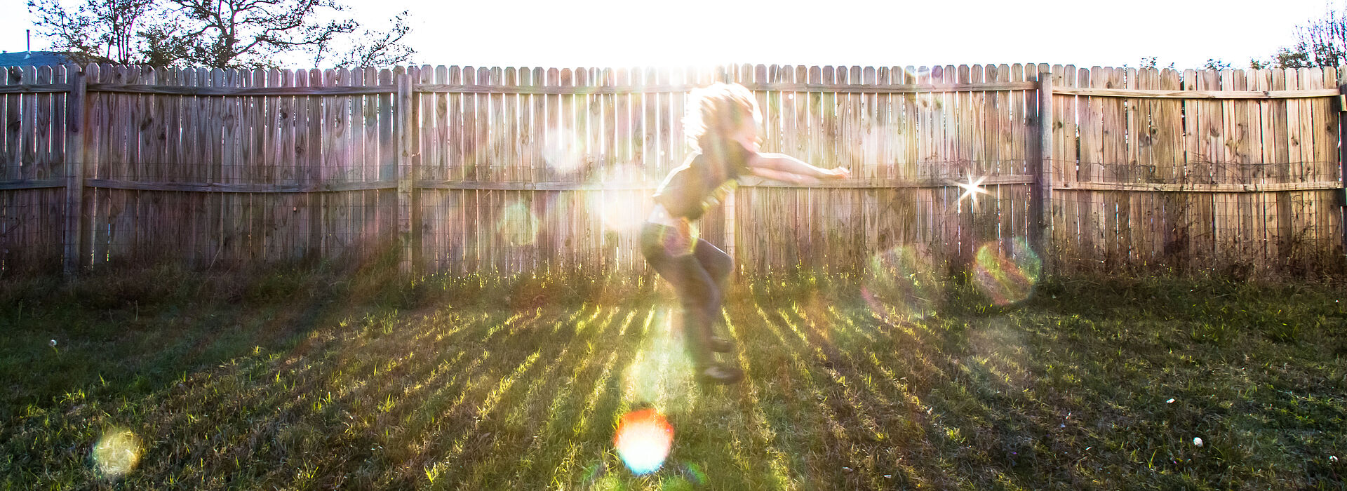 Kind vor einem Gartenzaun, spielt als würde es reiten. Gegenlicht.