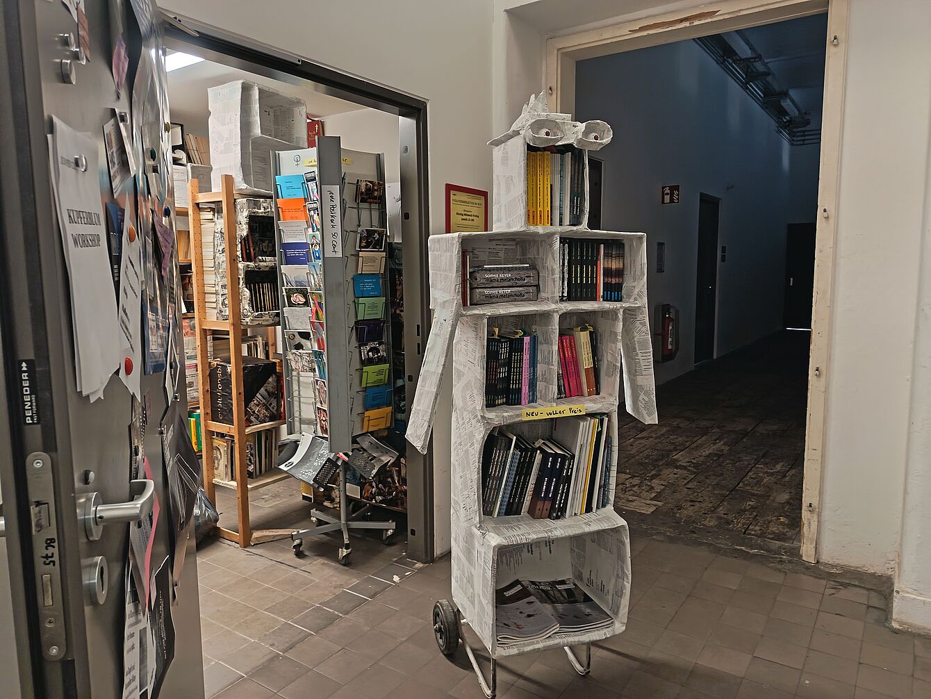 Mobiles Bücherregal in Form eines Roboters mit Brille aus Papier und Pappe in einem Flur vor einem Raum mit Regalen und Stühlen.