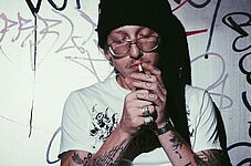 Edo Saiya am 15.4.2026 im WUK Wien - Pressefoto von Edo Saiya, der mit Mütze aus, an Wand angelehnt sitzend eine Zigarette anzündet