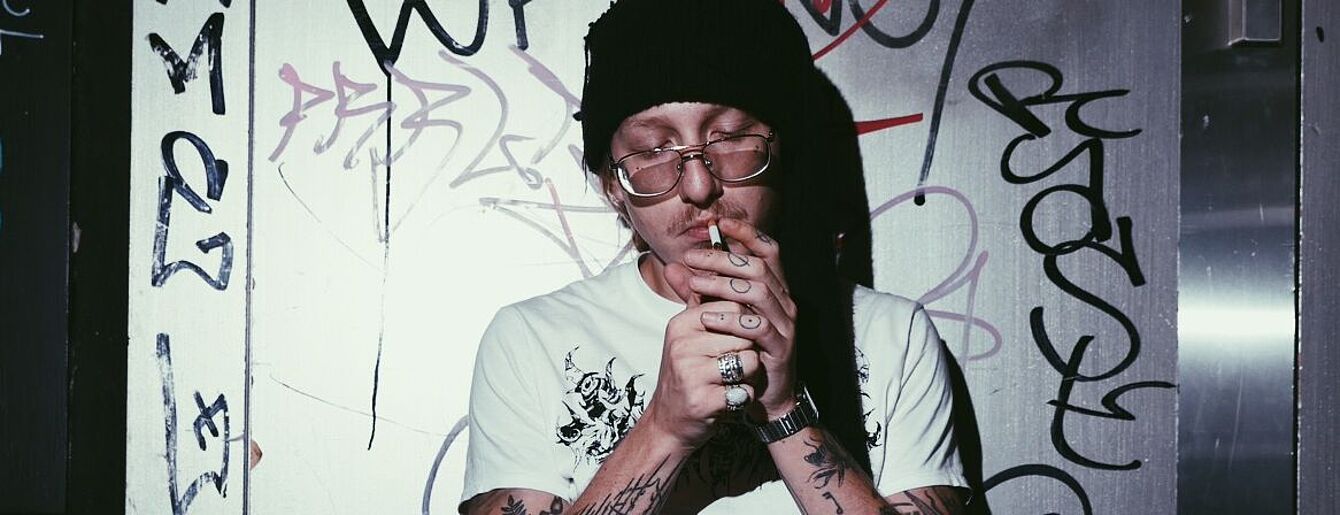Edo Saiya am 15.4.2026 im WUK Wien - Pressefoto von Edo Saiya, der mit Mütze aus, an Wand angelehnt sitzend eine Zigarette anzündet