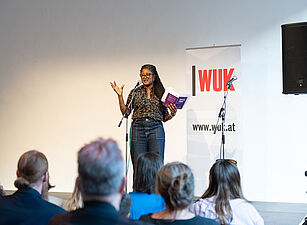 Frau mit Brille und Zöpfen steht vor Publikum und hält ein Buch in der Hand, im Hintergrund Banner mit Schriftzug 'WUK' und Webseite