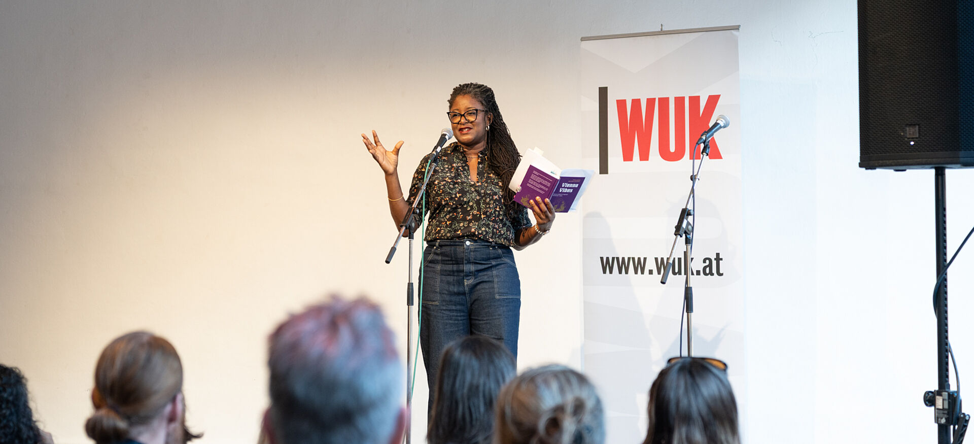 Frau mit Brille und Zöpfen steht vor Publikum und hält ein Buch in der Hand, im Hintergrund Banner mit Schriftzug 'WUK' und Webseite
