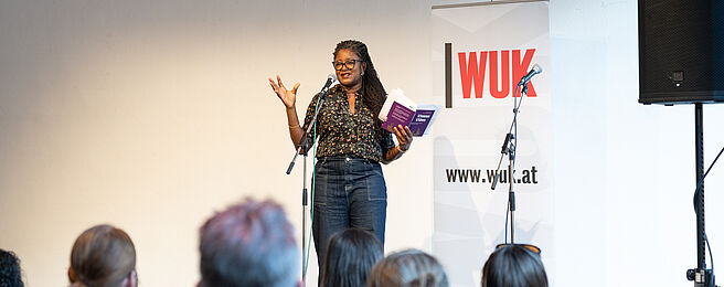 Frau mit Brille und Zöpfen steht vor Publikum und hält ein Buch in der Hand, im Hintergrund Banner mit Schriftzug 'WUK' und Webseite