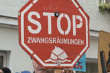 Weiblich und männlich gelesene Demonstrant*innen halten rote Stoppschilder mit der Aufschrift 'STOP ZWANGSRÄUMUNGEN' vor einem weißen Gebäude mit Fenstern.