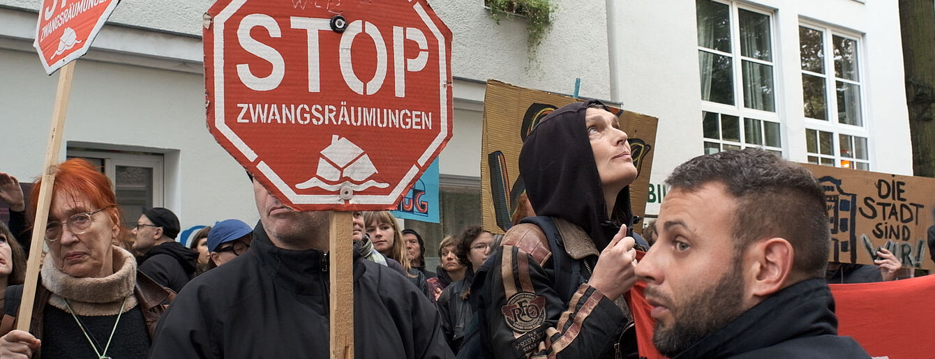 Weiblich und männlich gelesene Demonstrant*innen halten rote Stoppschilder mit der Aufschrift 'STOP ZWANGSRÄUMUNGEN' vor einem weißen Gebäude mit Fenstern.