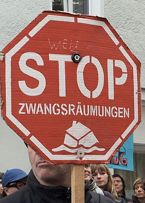 Weiblich und männlich gelesene Demonstrant*innen halten rote Stoppschilder mit der Aufschrift 'STOP ZWANGSRÄUMUNGEN' vor einem weißen Gebäude mit Fenstern.
