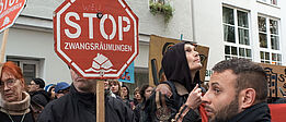 Weiblich und männlich gelesene Demonstrant*innen halten rote Stoppschilder mit der Aufschrift 'STOP ZWANGSRÄUMUNGEN' vor einem weißen Gebäude mit Fenstern.