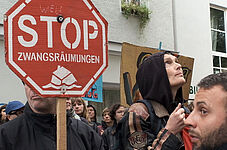 Weiblich und männlich gelesene Demonstrant*innen halten rote Stoppschilder mit der Aufschrift 'STOP ZWANGSRÄUMUNGEN' vor einem weißen Gebäude mit Fenstern.