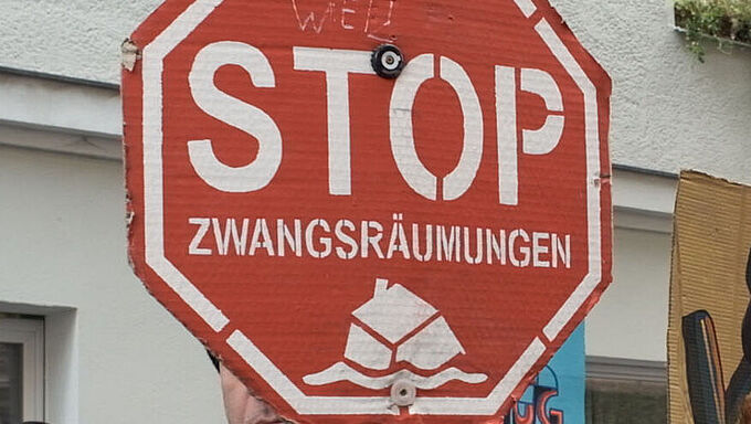 Weiblich und männlich gelesene Demonstrant*innen halten rote Stoppschilder mit der Aufschrift 'STOP ZWANGSRÄUMUNGEN' vor einem weißen Gebäude mit Fenstern.