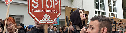 Weiblich und männlich gelesene Demonstrant*innen halten rote Stoppschilder mit der Aufschrift 'STOP ZWANGSRÄUMUNGEN' vor einem weißen Gebäude mit Fenstern.