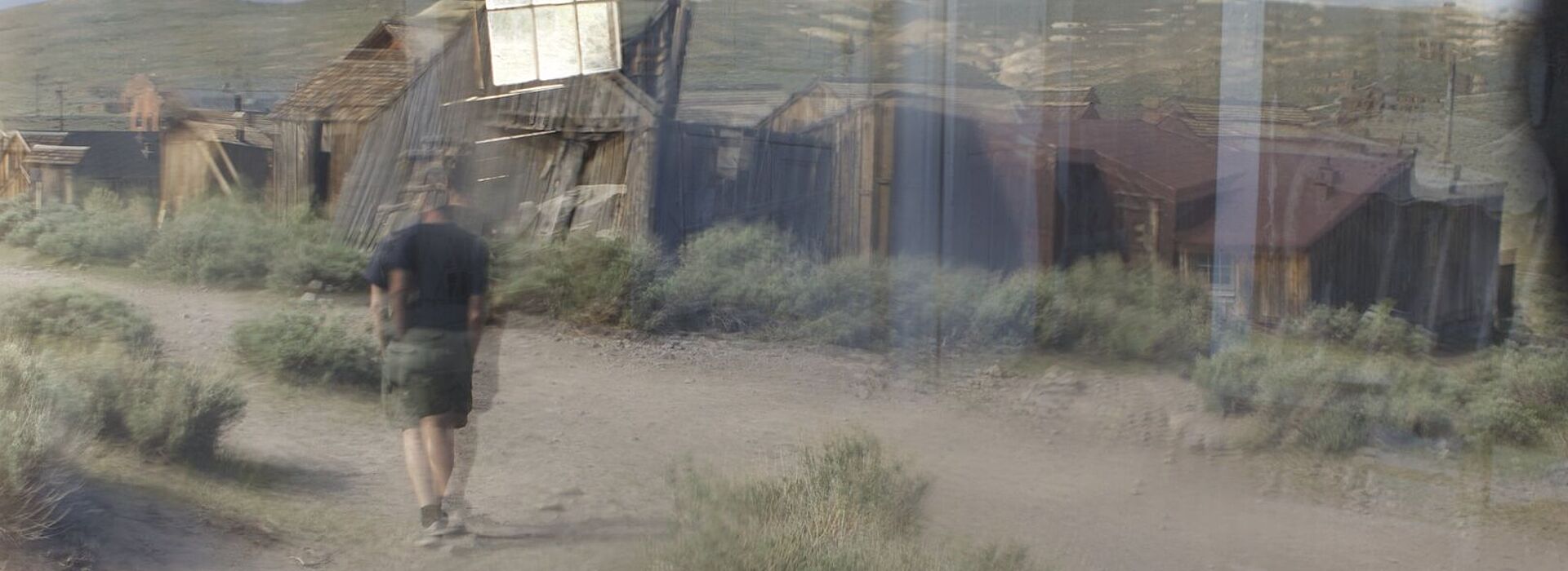 Verschwommene Fotografie, ein Mann in Freizeitkleidung geht auf einem Schotterweg auf hölzene Baracken zu.
