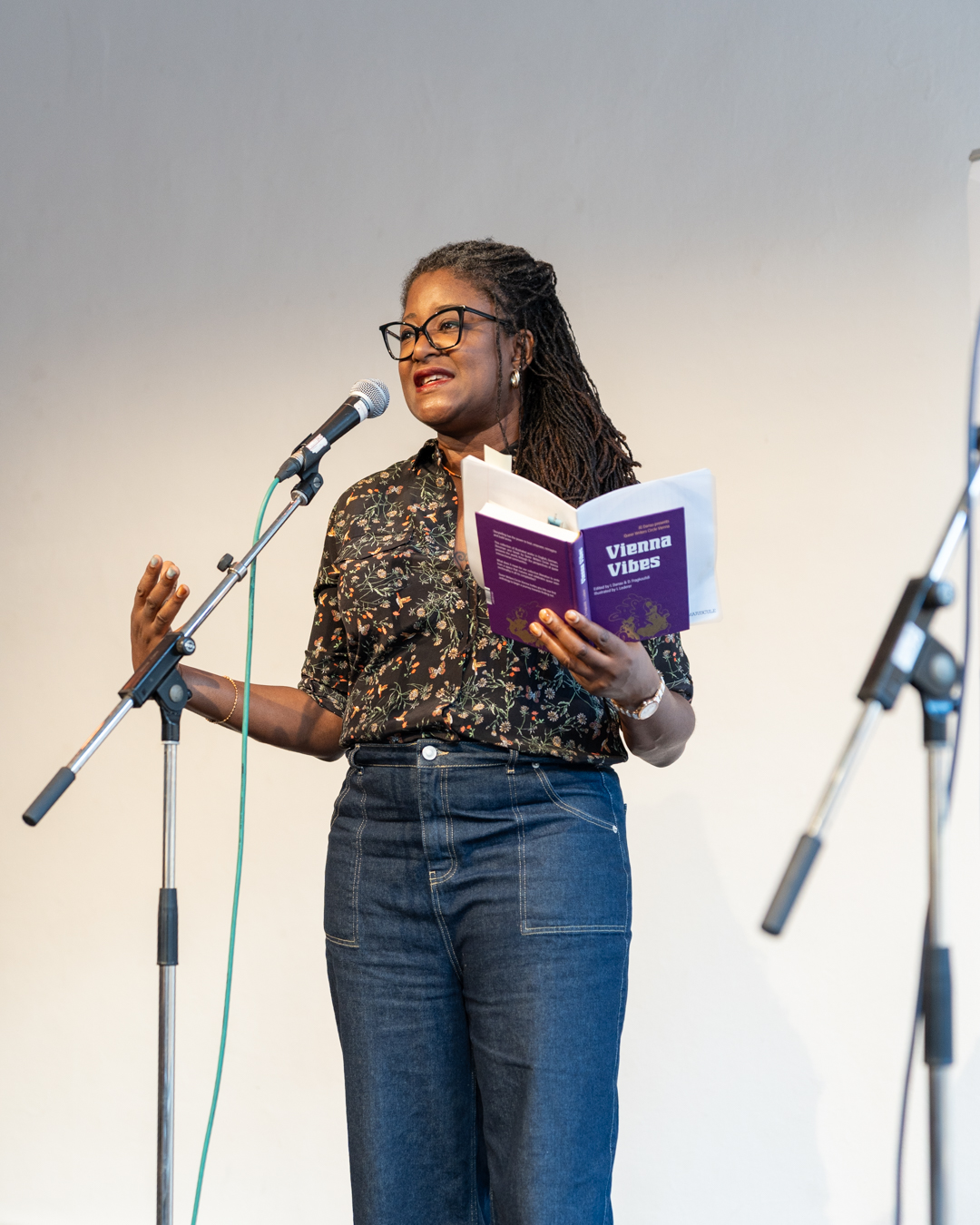 Eine Frau mit Brille und gemustertem Hemd steht vor einem Mikrofon und liest aus einem Buch mit dem Titel 'Vienna Vibes'