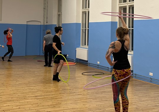 Mehrere Personen in einem Raum mit Holzboden drehen Hula-Hoop-Reifen um ihre Körper