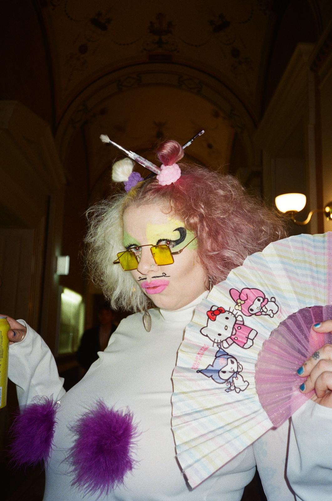 Person mit zweifarbigem lockigem Haar, gelben Brillengläsern, pinkem Lippenstift, hält einen Fächer mit Hello-Kitty-Motiven und trägt ein weißes Oberteil mit lila Pelzbesatz