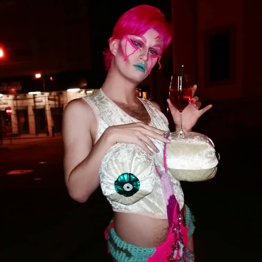Person mit pinkem Haar, auffälligem Make-up und grünem Lippenstift hält ein Glas Sekt und trägt ein weißes, gepolstertes Oberteil mit bunten Applikationen